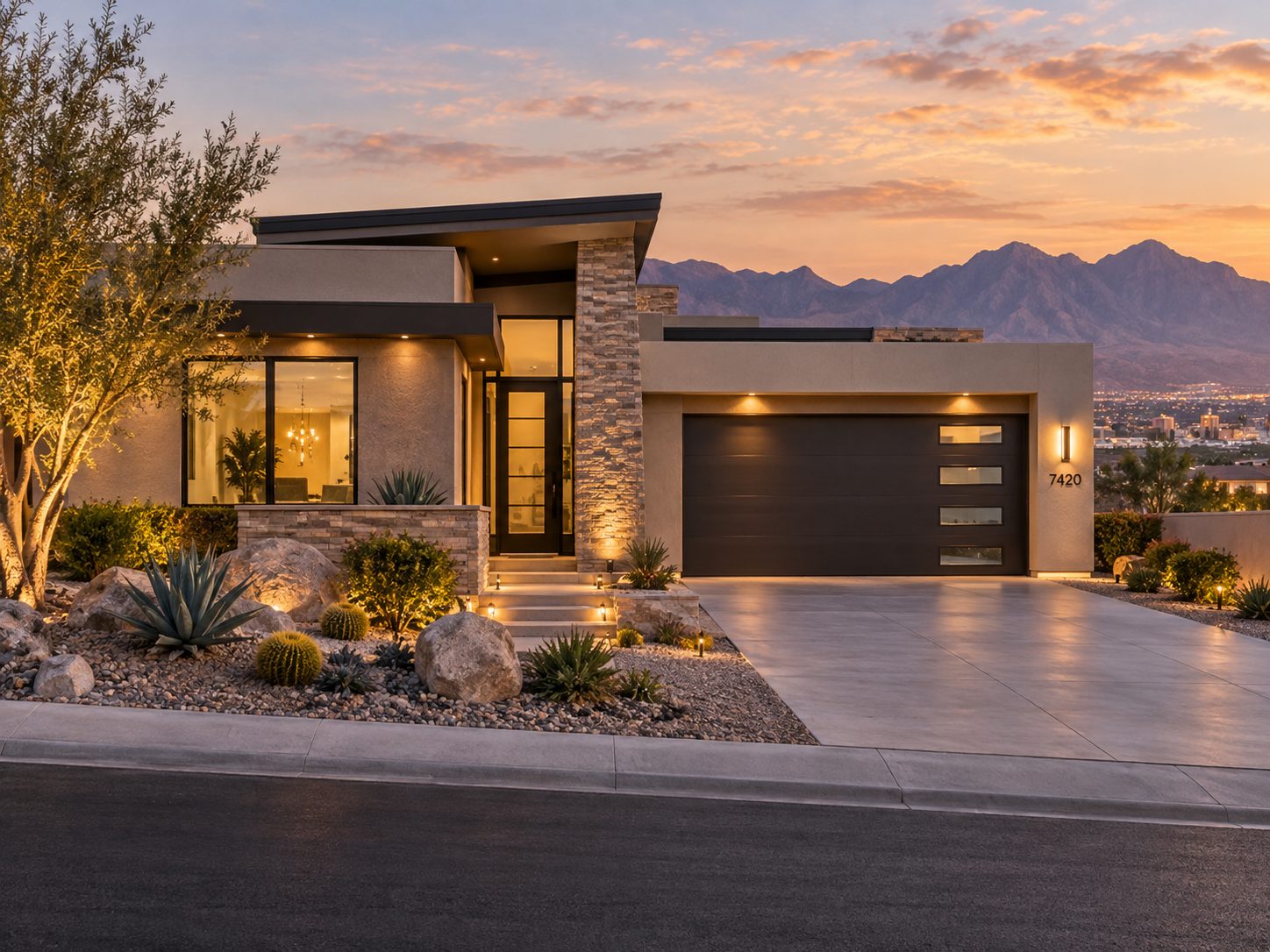 Modern desert home with stone accents and dark garage door lit at dusk, mountains beyond